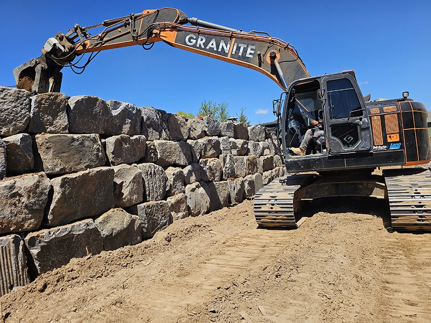 Equipment Excavator Placing Granite Boulders To Build A Retaining Wall | Granite Companies MN